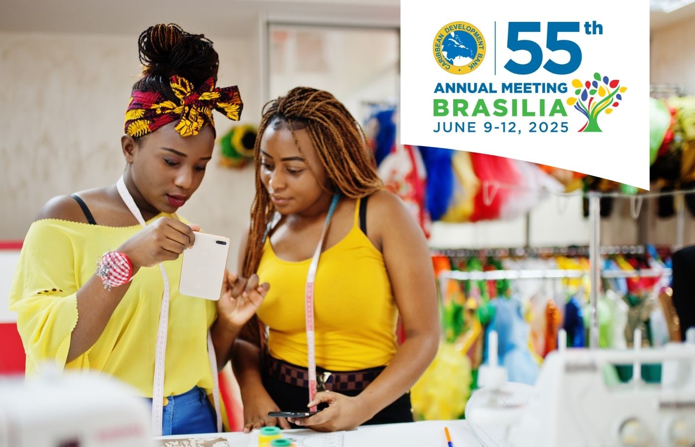 The image shows two women in a brightly lit fashion workshop. They are focused on a smartphone, seemingly discussing something related to their work. Both are wearing vibrant yellow outfits, with measuring tapes around their necks, and are surrounded by colorful fabric designs and sewing equipment in the background.