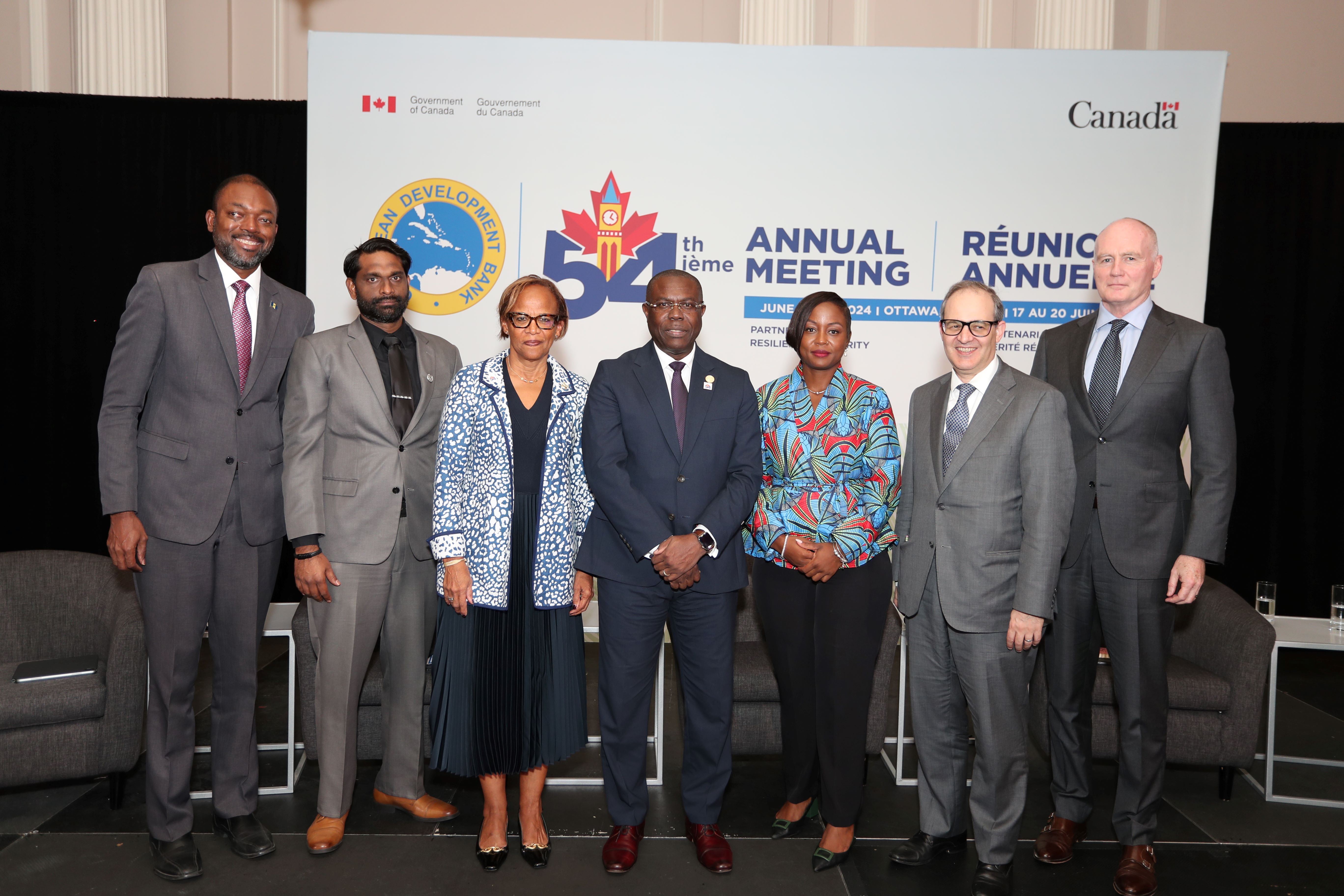 Seven people stand on a stage in front of a backdrop of the CDB's Annual Meeting.