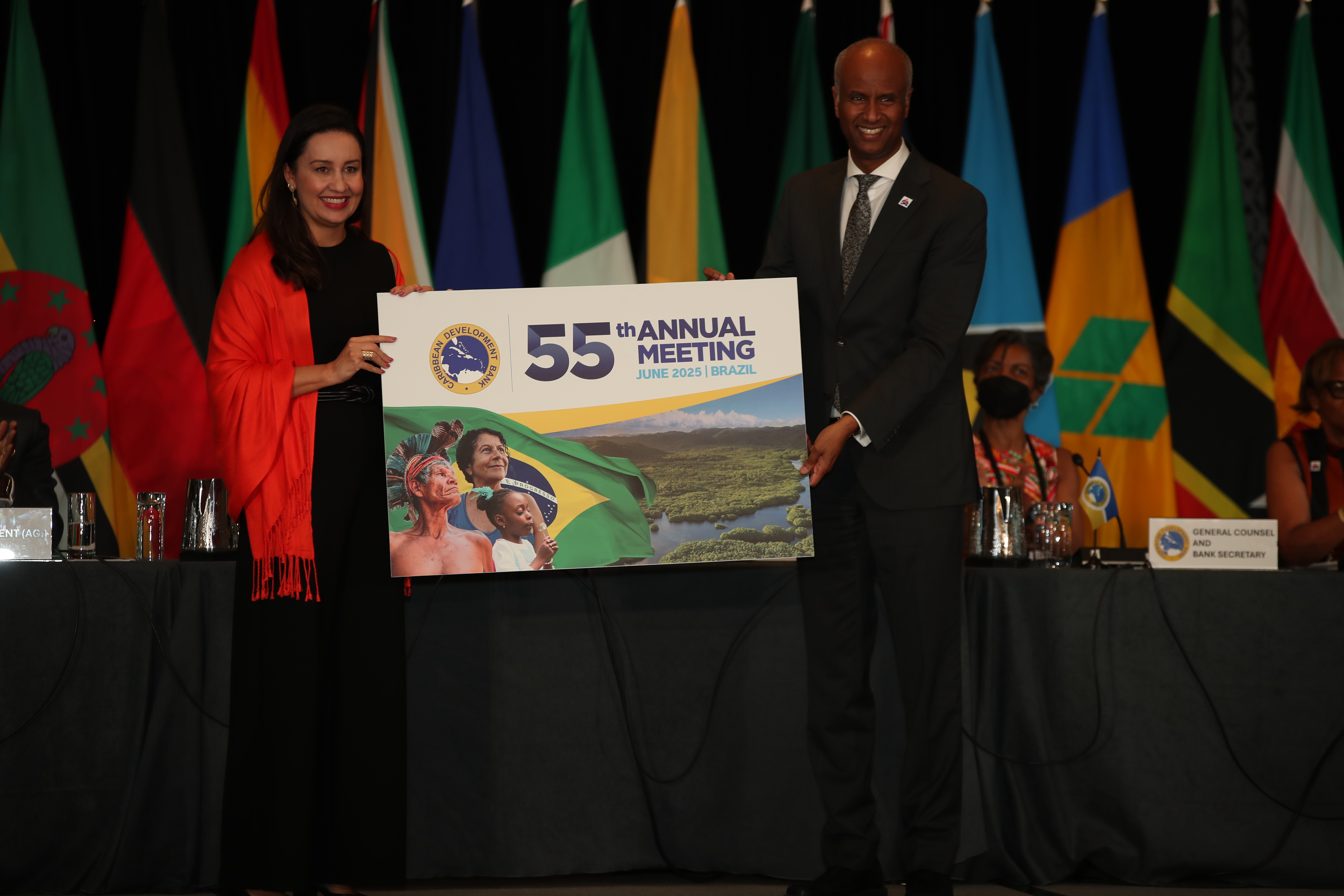 A woman and man stand on a platform in front of several flags of CDB member countries. The man is handing over a card to the woman with the words 55th Annual Meeting 