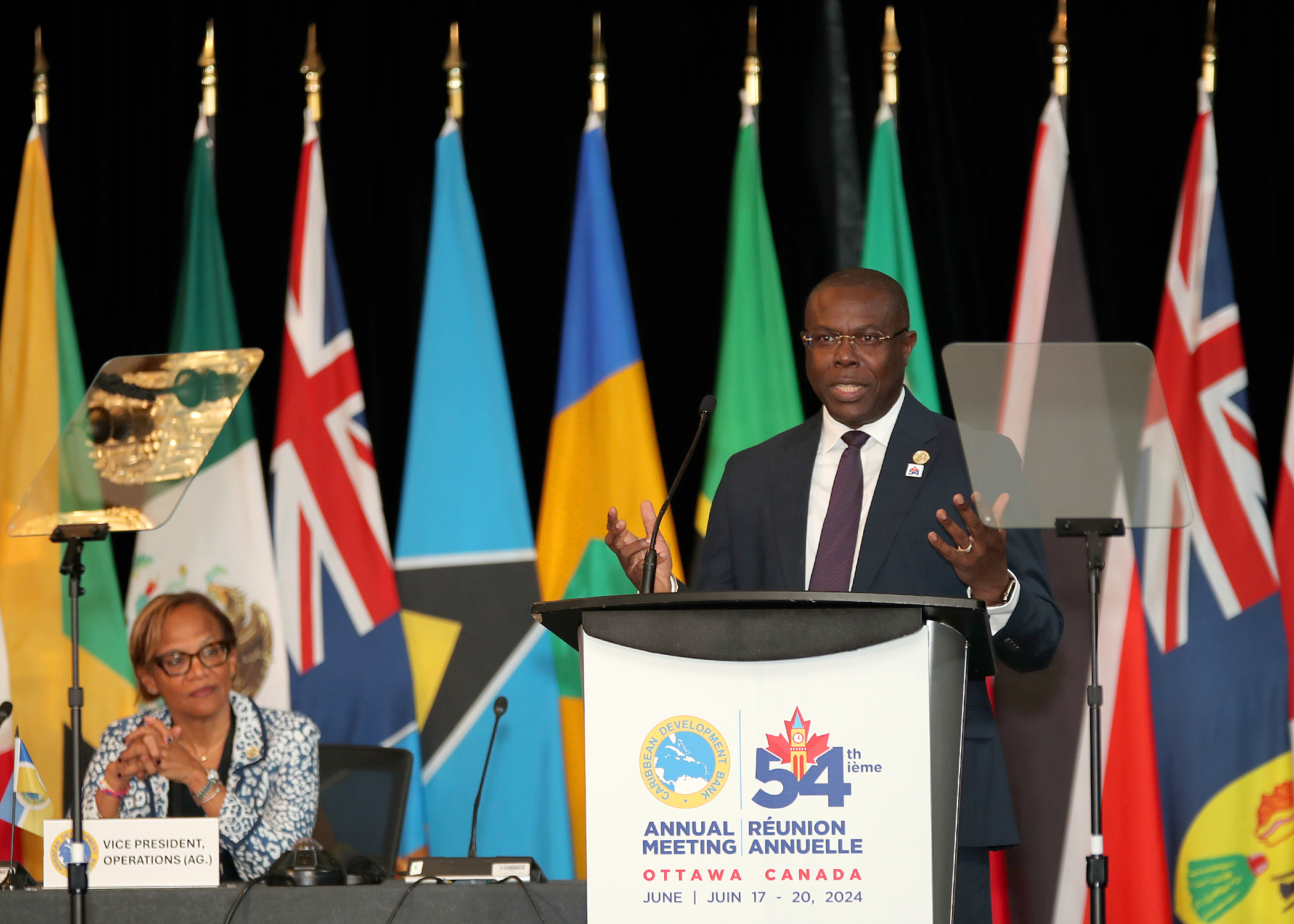 A man stands on a platform behind a lectern with the logo and words Caribbean Development Bank 54th with several flags behind him