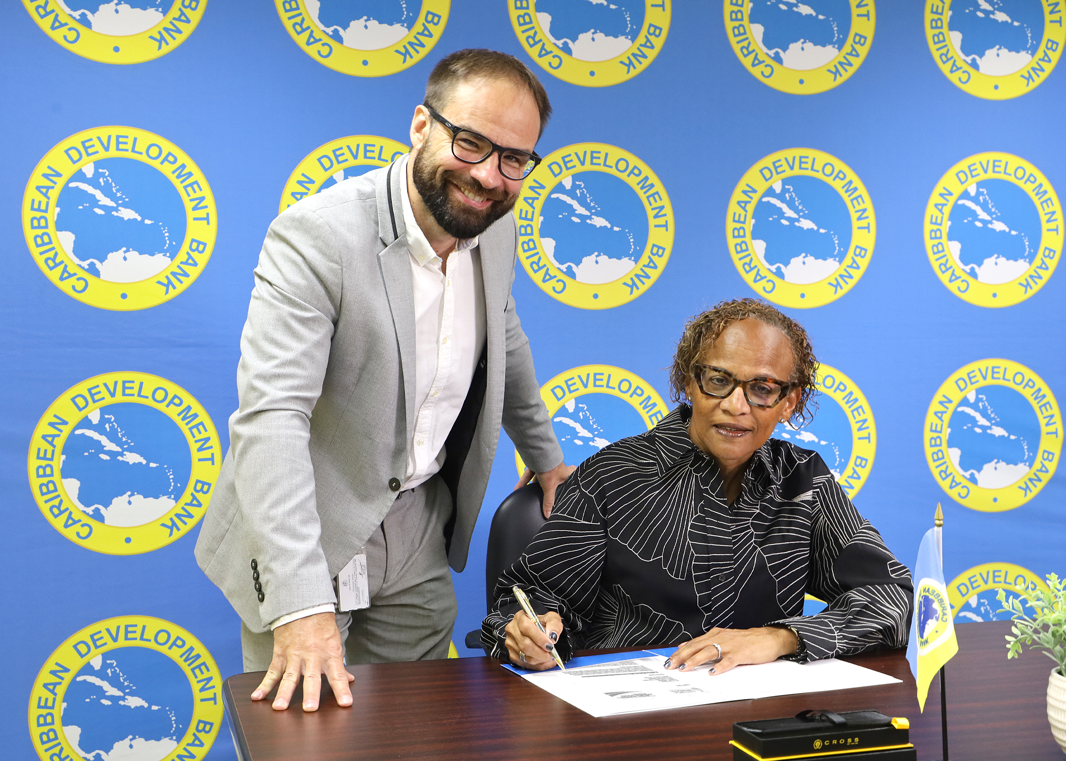 Lady seated in black and white patterned with pen signing and man in tan suit leaning over table and looking forward