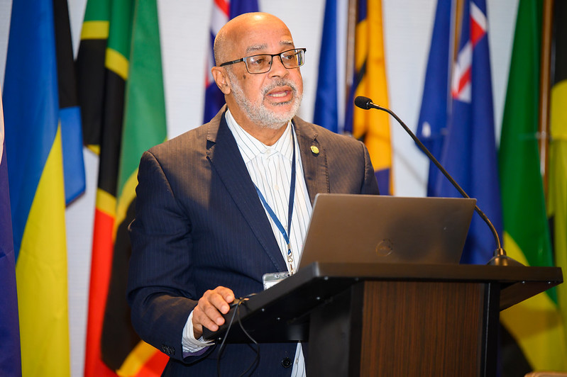 Dr. Didacus Jules stands behind a lectern with several Caribbean country flags hoisted behind him