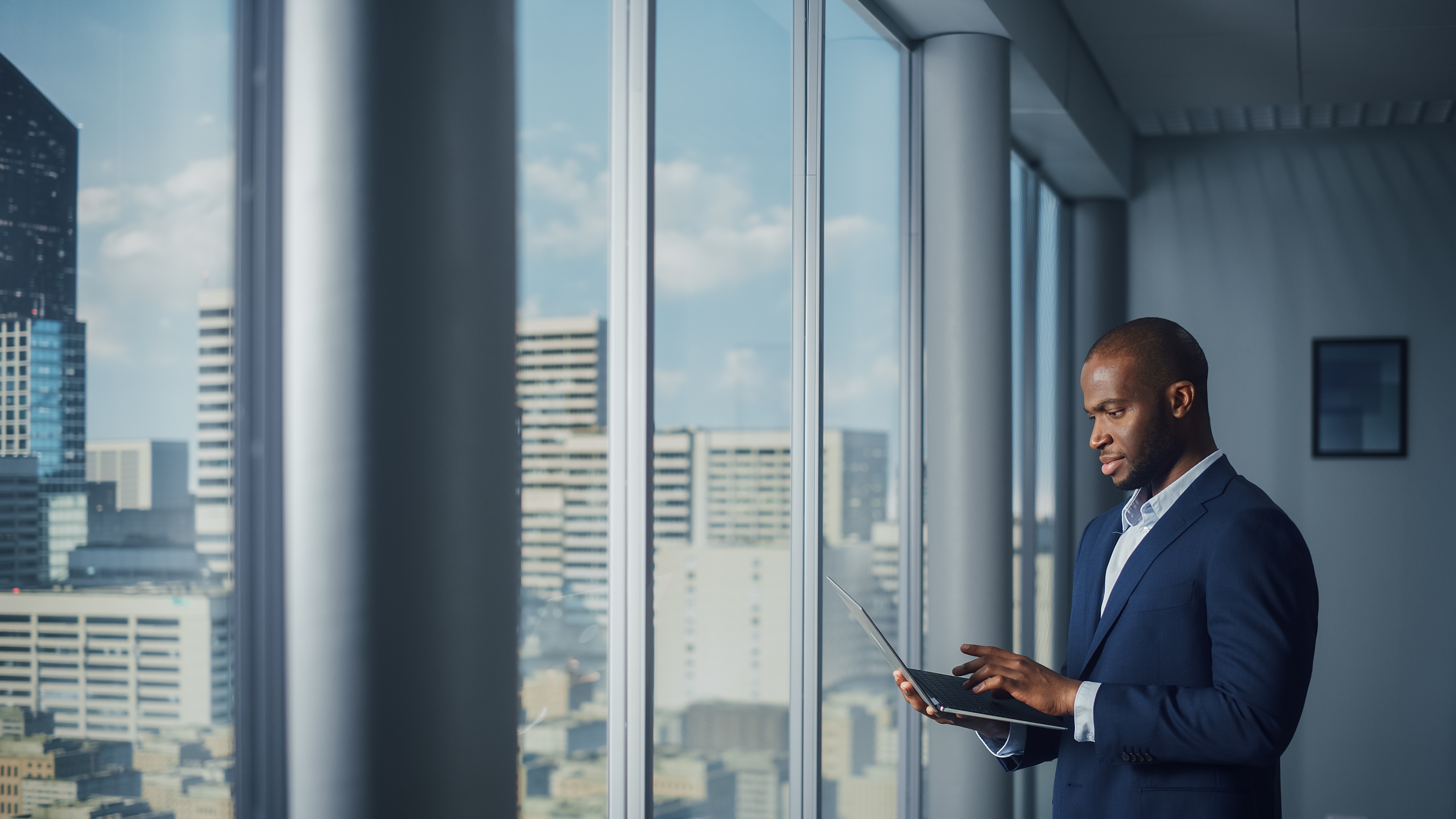 Thoughtful Black Businessman in a Tailored Suit Using Laptop while Standing in Office Near Window on Big City.