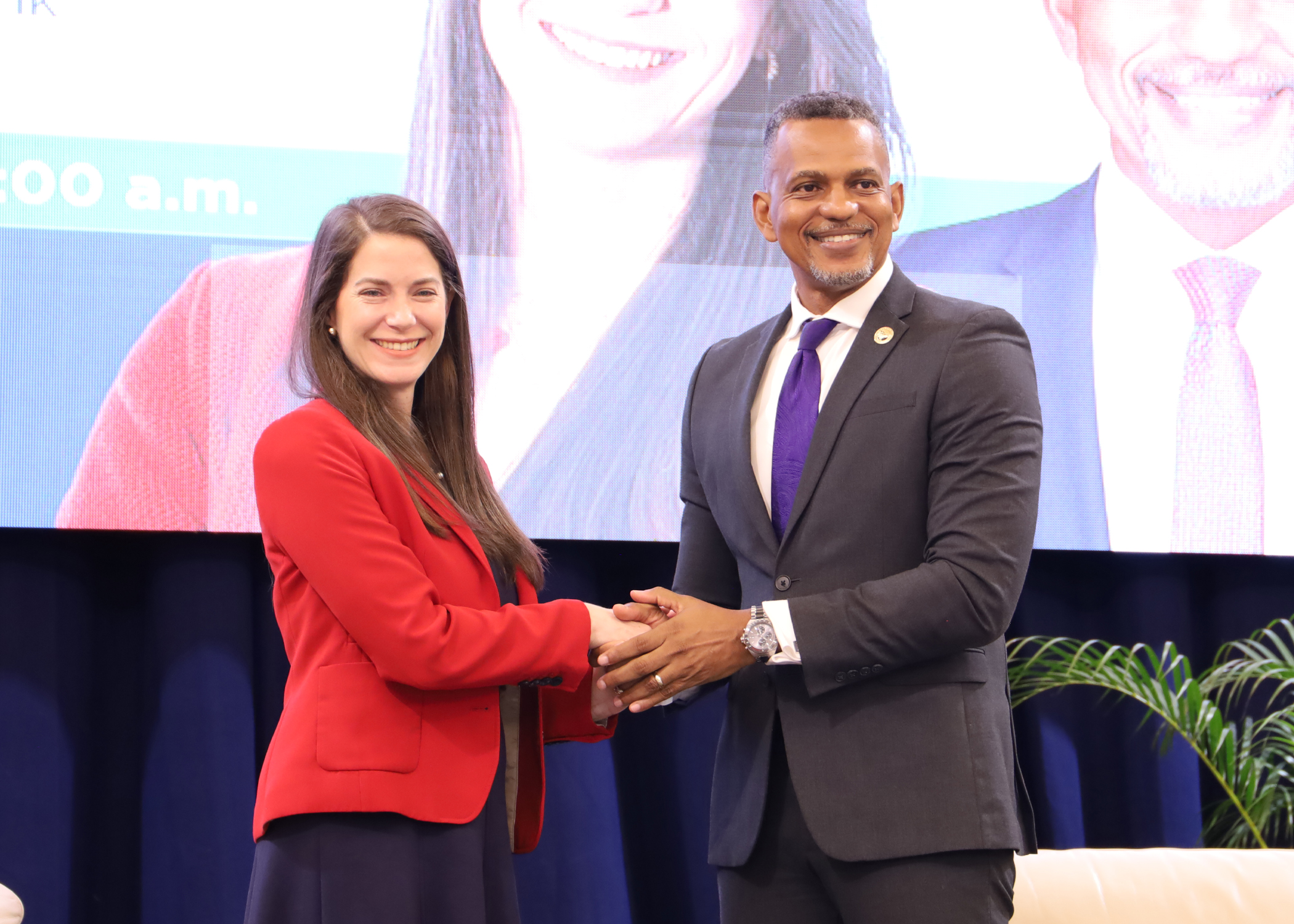 A woman and a man in formal attire are shaking hands and smiling for the camera against a stage backdrop, with their images displayed on a screen behind them