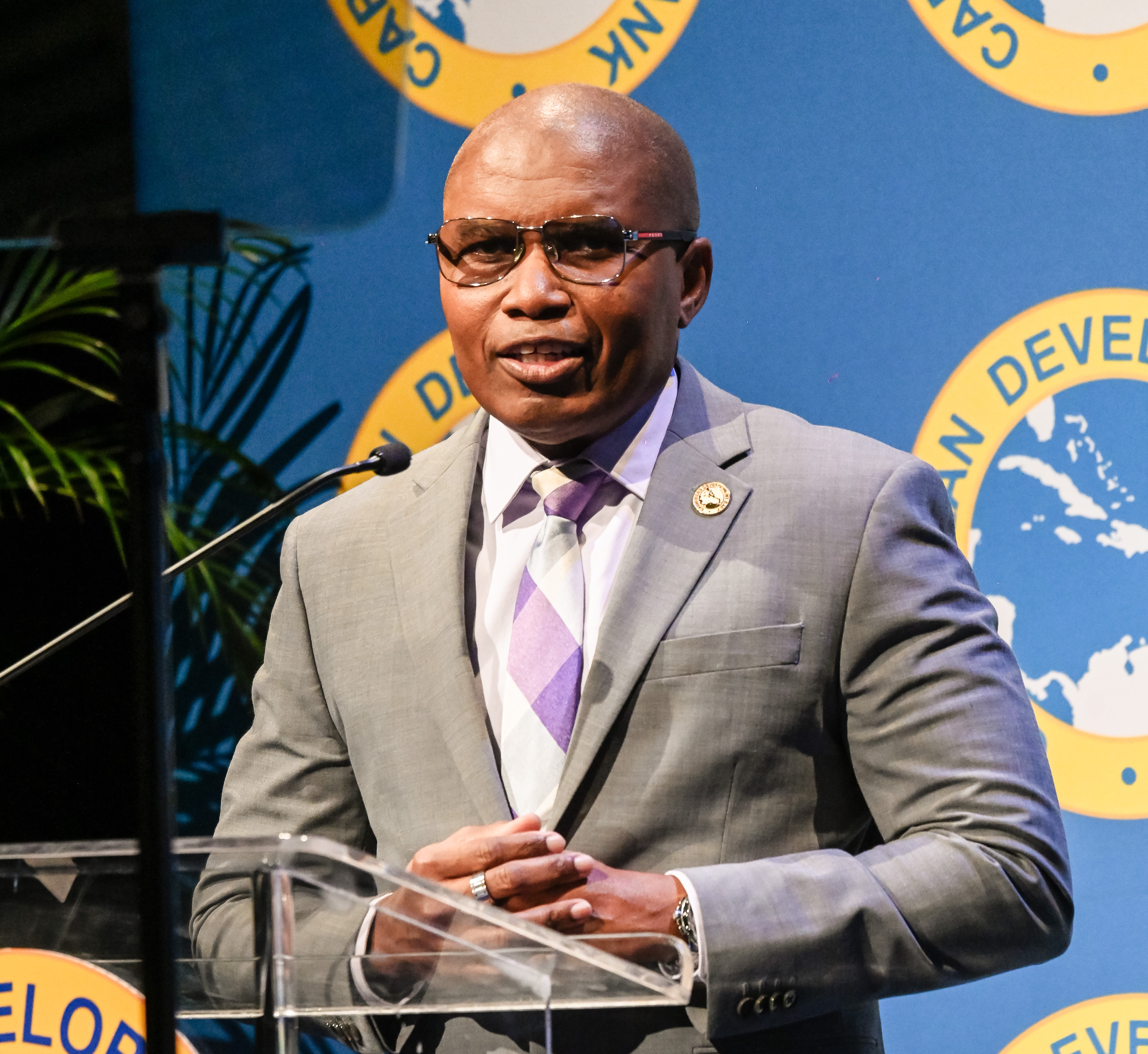 CDB Director of Economics, Mr. Ian Durant dressed in a grey suit with a pink and grey tie speaks at a podium in front of a backdrop displaying the logo of the Caribbean Development Bank.