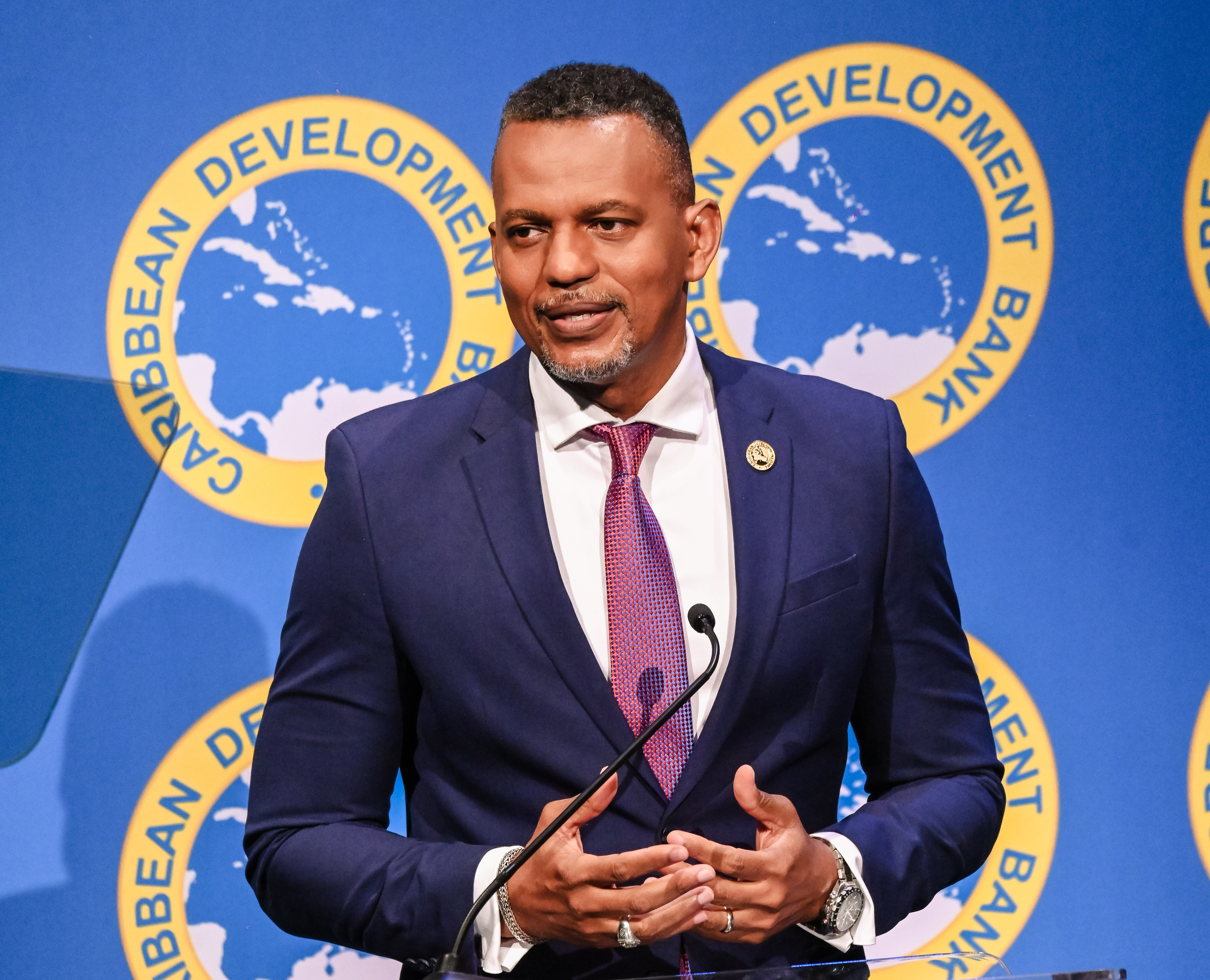 CDB President, Mr. Daniel Best dressed in a navy suit with a red tie speaks at a podium in front of a backdrop displaying the logo of the Caribbean Development Bank.