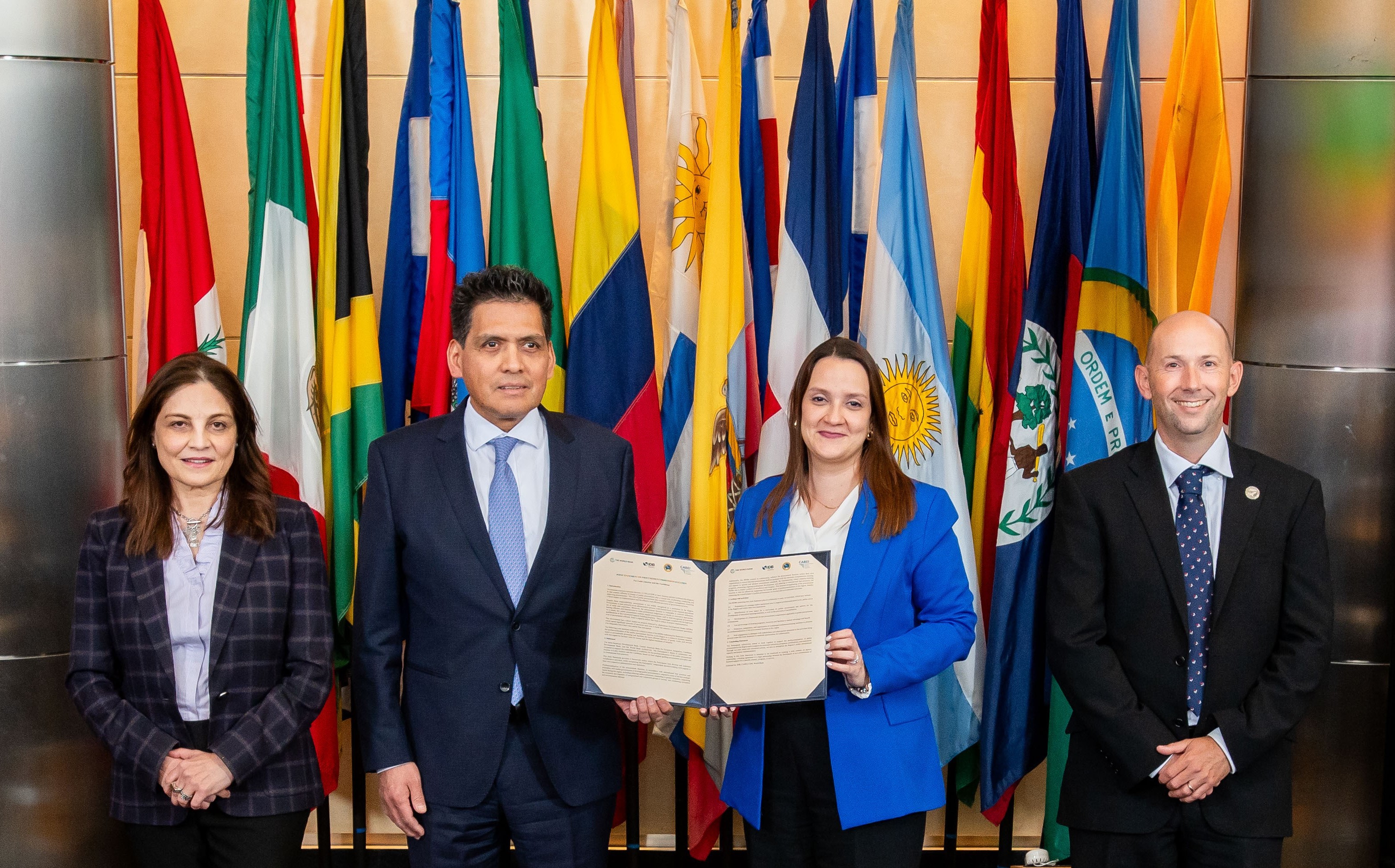 Four individuals stand in front of a row of national flags, with two in the center holding a signed document. The individuals are professionally dressed, and the flags represent various countries in the background