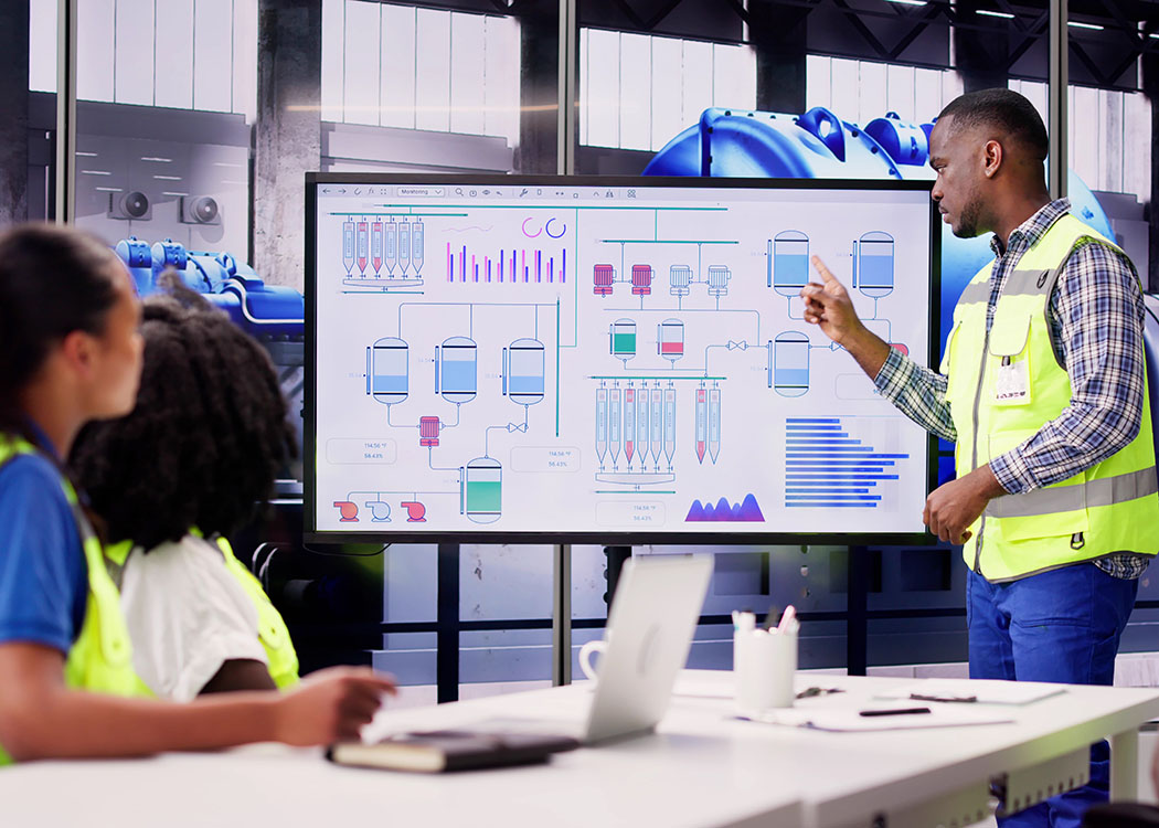 man in reflective vest standing in front of white board with colourful charts pointing to one of the graphs as two female colleagues whi are seated look on