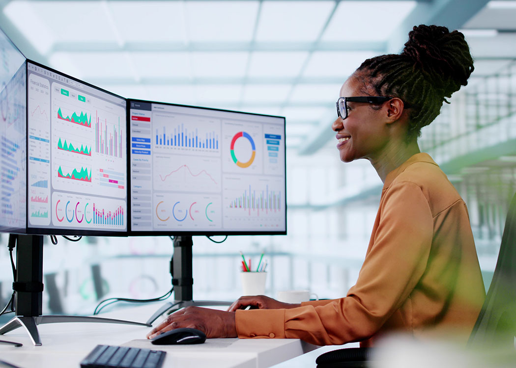 A professional woman seated at a desk in a modern office, analyzing data displayed on dual monitors. The screens show colorful charts, graphs, and financial statistics. Natural light filters through large windows in the background.