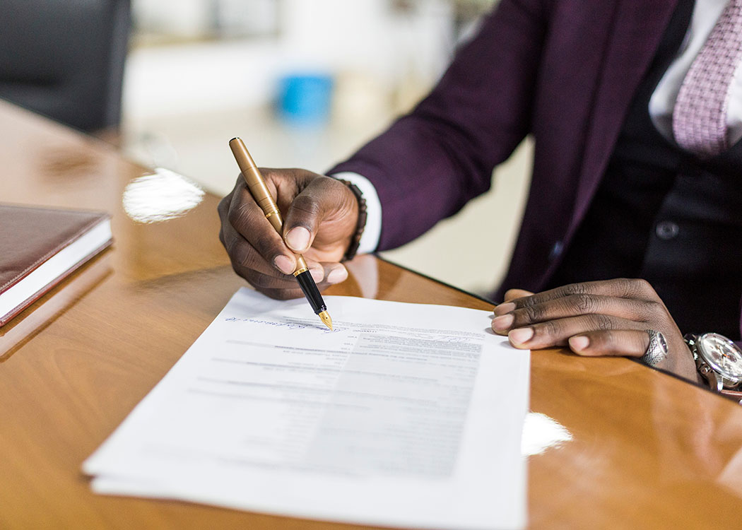 Close-up image of a person's hand signing a document with a gold fountain pen on a polished wooden desk. A closed leather-bound notebook and other office items are visible in the background