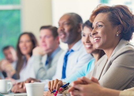 Image of a diverse group of professionals seated at a workshop or training session. They are engaged and smiling, with notebooks and coffee cups visible on the table. Natural light streams through a window in the background.