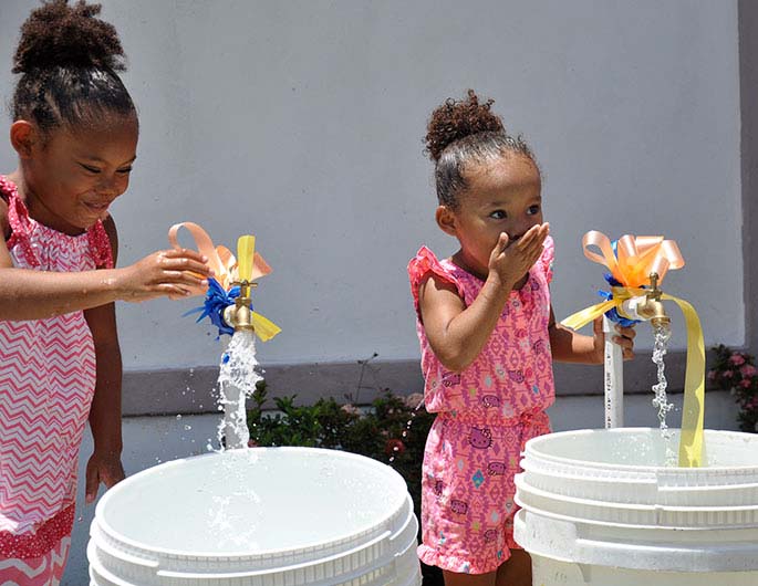 two little girls with cupped hands drinking water from an external pipe