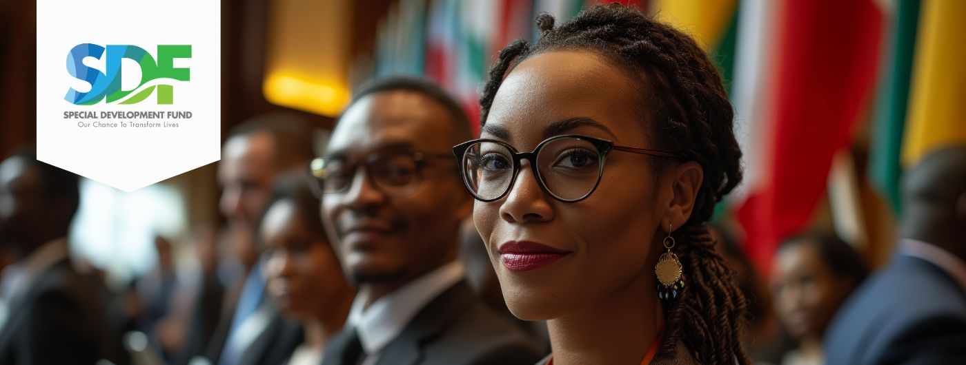 Image of a confident woman with glasses and braided hair attending a formal event, seated among diverse individuals in business attire. She is in focus, with international flags blurred in the background. The logo of the Special Development Fund (SDF) and the tagline 'Our Chance to Transform Lives' are displayed in the upper left corner.