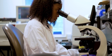 young black girl in white coat, seated and looking into a microscope