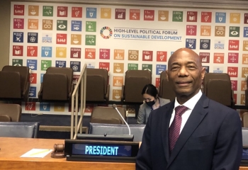 gentleman in dark suit with white shirt and red tie standing in front of a backdrop with the SDG icons