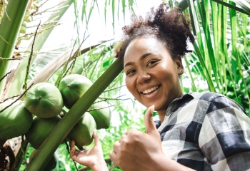 Smiling young black girl standing by a coconut tree giving a thumbs up while looking into the camera