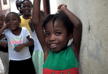 black girl with plaited hair looking into the camera while leaning against the wall, as a group of smiling children her age in the background