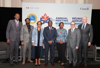 Seven people stand on a stage in front of a backdrop of the CDB's Annual Meeting.