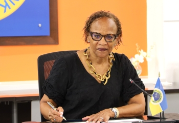 Bespectacled black woman in black dress seated looking on camera holding pen