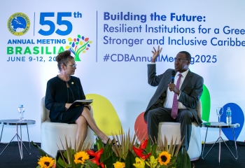 Dr. Clarke and Ms. Poire are seated on a stage at the 55th Annual Meeting of the Caribbean Development Bank in Brasília, Brazil. On the left, a woman holds a notebook, attentively listening, while on the right, a man gestures while speaking into a microphone. A colorful backdrop displays the event's theme, "Building the Future: Resilient Institutions for a Greener, Stronger, and Inclusive Caribbean."