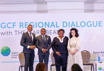 A photo from the GCF Regional Dialogue with the Caribbean, held in Saint Kitts and Nevis from March 17–20, 2025. Four people are standing on stage in front of a banner displaying the event name and Green Climate Fund logo. From left to right: a man in a dark suit, a man in a navy suit, a woman in a navy pantsuit with a pin, and a woman in a cream dress.