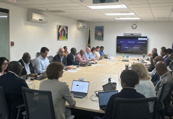 A group of professionals from the Caribbean Development Bank and the World Bank are seated around a large rectangular conference table in a modern meeting room. Most participants are using laptops, and several are engaged in discussion. On the wall ahead is a large screen displaying names of remote attendees, indicating a hybrid meeting setup. Flag of Jamaica is in the left corner.