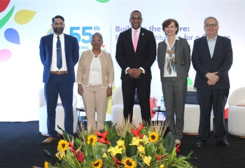 A group of five individuals stands on a stage at the 55th Annual Meeting of the Caribbean Development Bank, with the event's theme "Building the Future: Resilient Institutions for a Greener, Stronger, and Inclusive Caribbean" displayed on the backdrop. The individuals, representing a mix of formal attire, are positioned in front of a vibrant floral arrangement featuring sunflowers and tropical plants. The event logo is prominently visible in the background.