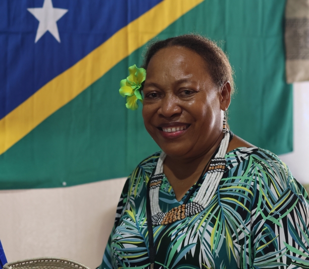 Pacific woman with yellow flower in her hair and green yellow black and white dress with multicolored beaded necklace